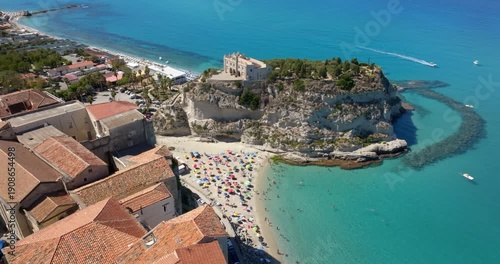 Aerial view of Sanctuary of Santa Maria dell'Isola in Tropea, Calabria, Italy. The ancient church stands on a rocky promontory, overlooking a crowded sandy beach filled with colorful sun umbrellas.