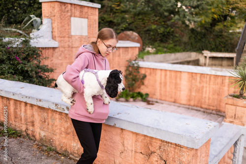 Teenage blonde Slavic girl carrying her Russian spaniel dog along a stone wall in Montenegro