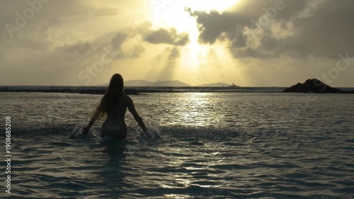 In this slow motion 4K video from Seychelles, a woman enjoys her tropical vacation, jumping happily in the warm ocean water at sunset. With her arms raised and water splashing around her, she celebrat
