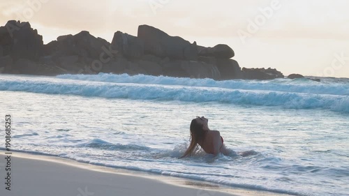 Slow motion 4K video of a woman relaxing in the gentle waves on a Seychelles beach during sunset. She leans back in the water, enjoying a peaceful tropical vacation surrounded by turquoise sea and dra