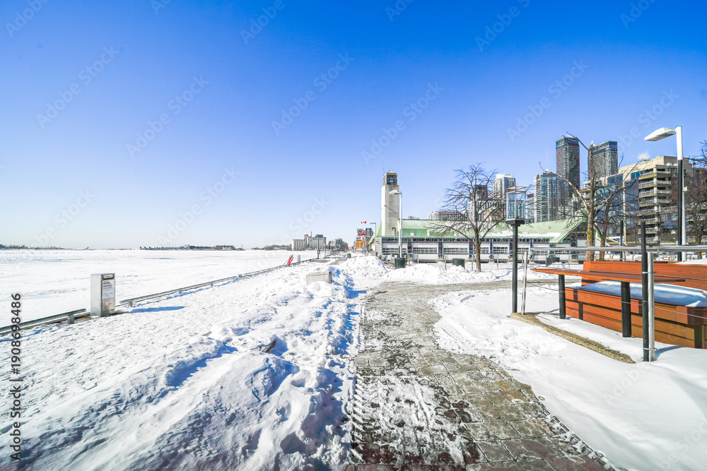 Obraz premium Toronto harbourfront centre blanketed in snow under a clear blue sky, overlooking the frozen expanse of lake ontario with skyline, park paths and serene winter cityscape