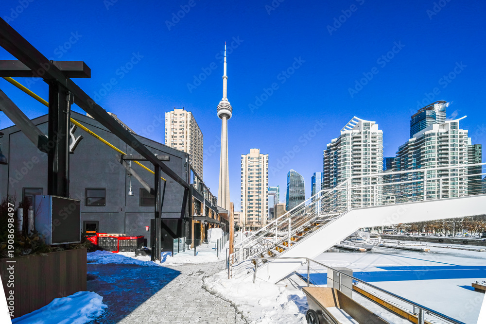 Fototapeta premium Toronto cityscape showcasing a prominent landmark tower rising above urban buildings and a waterfront area covered in snow on a bright, clear blue sky day