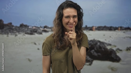 Young hispanic woman holds finger to lips for silence on beach near rocky shoreline under natural sunlight; mystery.