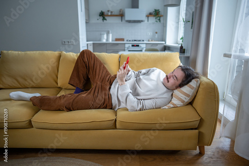 Schilderij op canvas Lazy bored man lying on sofa scrolling phone with social media, procrastinating, lack of motivation
