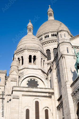 Low angle view of the white domes and intricate stone architecture of the Sacre Coeur Basilica in Montmartre, Paris, under a clear blue sky, famous French religious landmark and tourist attraction