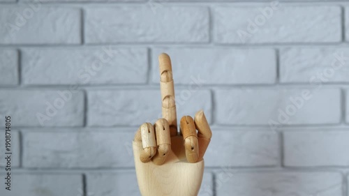 Wooden mannequin hand making obscene gesture. Articulated wooden hand model rising from the bottom of the frame to show an offensive middle finger gesture against a gray brick wall background