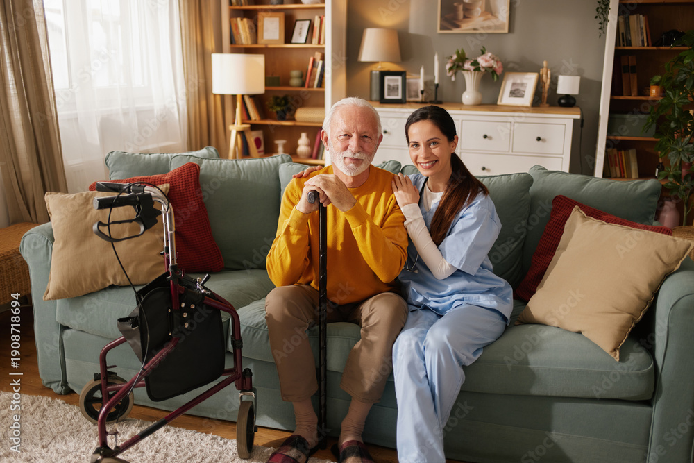 Obraz premium Nurse sits with elderly man on couch in living room. They talk while man holds cane. Walker is nearby. Room has bookshelves and soft light coming from window.
