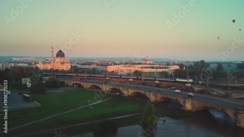 At dawn in Dresden's Old Town, a train crosses the bridge over the Elbe River, with the Yenidze Theater and Heinz-Steyer-Stadion in the background and a bird's-eye view of hot air balloons in the sky