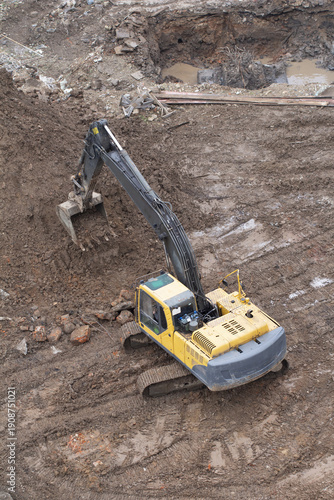 A yellow hydraulic excavator is digging into a pile of dirt on a muddy construction site. Concept of earthworks and site preparation.