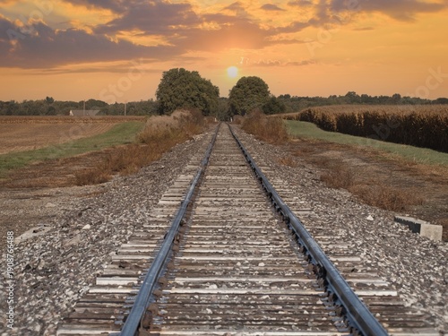 Sunset over rural railroad tracks flanked by cornfields, trees, and golden autumn grasses in scenic landscape