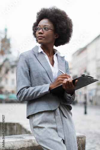 A professional African American woman in a suit and glasses is thoughtfully writing notes on a clipboard while standing outside in an urban setting