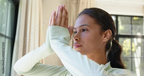 Adult African woman lifting arms, pressing palms into prayer near window in green top, meditating