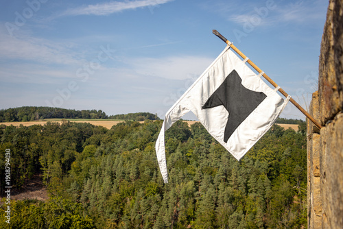 A white flag with a distinctive black emblem hangs on a wooden flagpole attached to a castle tower overlooking a dense forest