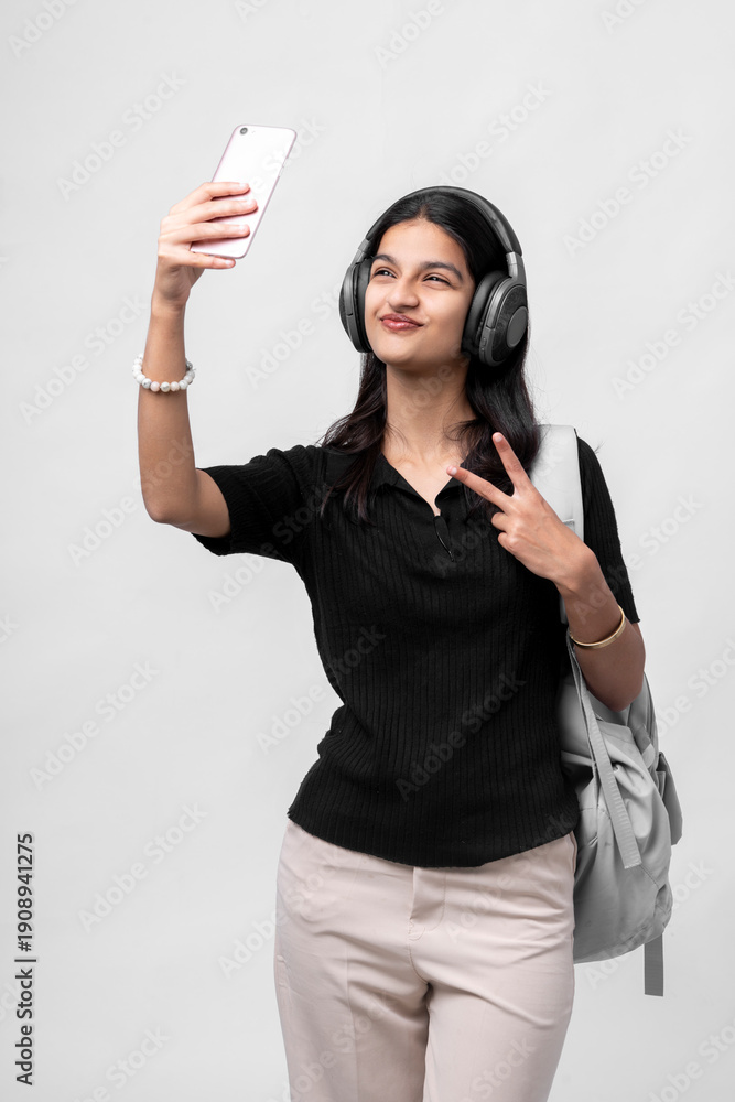 Naklejka premium young indian college girl with backpack and headphone taking selfie with smartphone