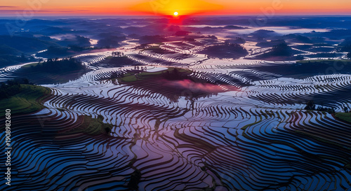 Fiery sunset glows above endless mirrored rice terraces, winding like ribbons across rolling hills in a breathtaking aerial landscape

