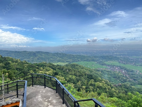 Panoramic high-angle view of Yogyakarta city and lush green rice fields under a blue sky. Beautiful landscape of Java, Indonesia, seen from a mountain hill during a clear day.