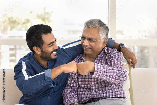 Happy young Indian man hugging and fist bumping older grandpa or great-grandfather seated together on couch, showing unity, understating, share support. Intergenerational family ties, mutual respect