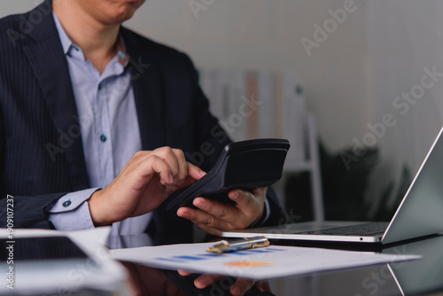 Close up businessman hands holding and using calculator to calculate financial data, tax and budget at office desk. Accountant working with laptop and business report documents.