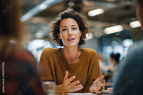 Caucasian businesswoman with short curly hair and mustard knit sweater explaining with open hands, animated and thoughtful in a bright startup coworking space