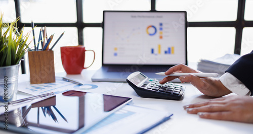 Focused businesswoman wearing glasses reads documents at a modern office desk, surrounded by a laptop, charts, and financial reports, exuding professionalism and concentration

