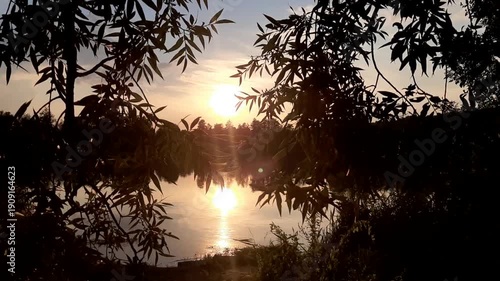 Golden sun sets behind forest silhouettes reflecting in calm lake water at dusk near Regensburg