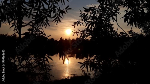 Golden sun sets behind forest silhouettes reflecting in calm lake water at dusk near Regensburg