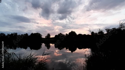 Golden sun sets behind forest silhouettes reflecting in calm lake water at dusk near Regensburg