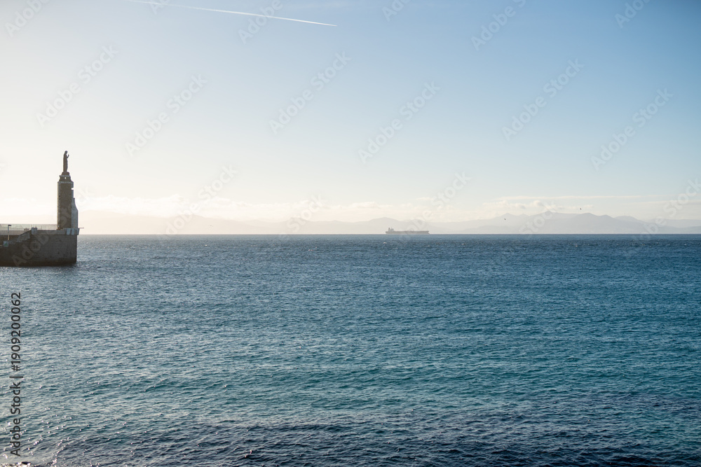 Fototapeta premium Wide ocean view with lighthouse statue on pier distant cargo ship and hazy mountains under soft sunlight creating tranquil maritime landscape with open water and horizon