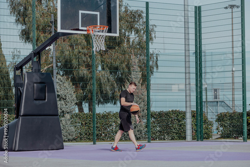 Young Hispanic man dribbles a basketball on an outdoor court. He wears a black shirt and shorts, with red sneakers. The background features a basketball hoop and greenery.