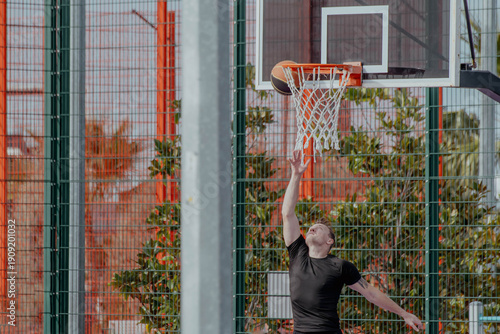 Young Caucasian man with short hair jumps to shoot a basketball at an outdoor court. Snow-capped mountains and city buildings are visible in the background.