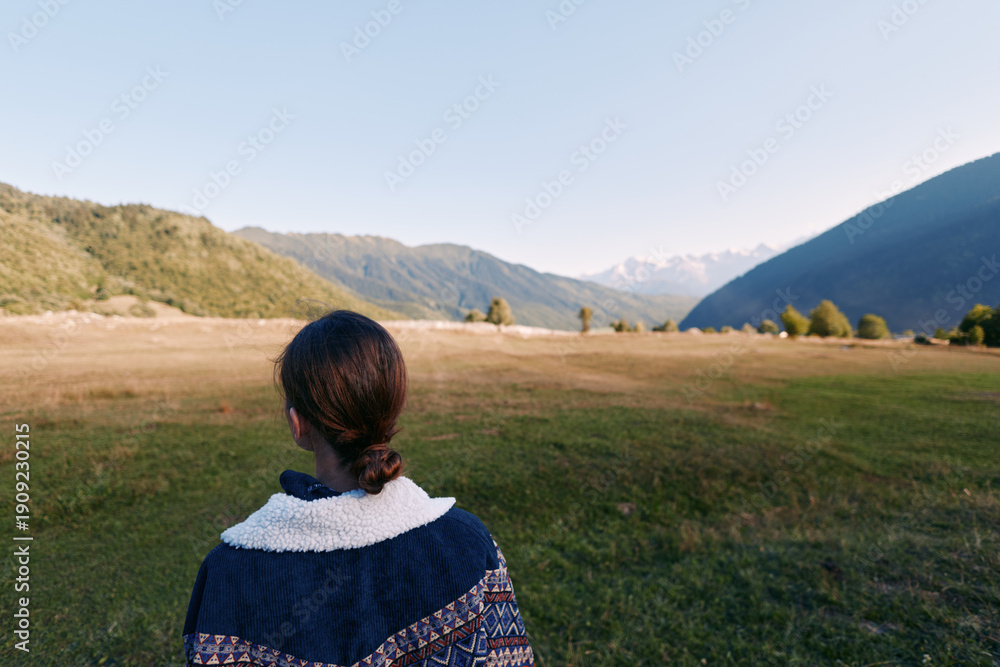 Fototapeta premium person woman mountains valley meadow nature back landscape in this photo: a woman seen from behind stands in a wide grassy meadow looking toward distant mountain valley under clear sky