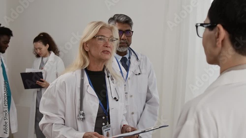 Over the shoulder shot of short-haired female health professional having discussion with mature doctor wearing stethoscope around her neck and holding clipboard, male specialist watching them