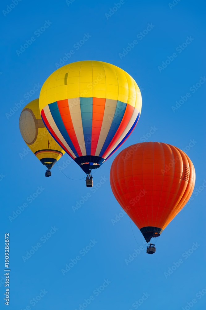 Obraz premium Three colorful hot air balloons with brightly colored envelopes and balloonists in the baskets hanging below on a sunny day in the Sauerland region (Germany), set against a cloudless blue sky. 