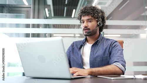 Young male corporate operator in headset working in customer support service on helpline telesale talking using laptop sitting in business office. The agent of the call center advises client online