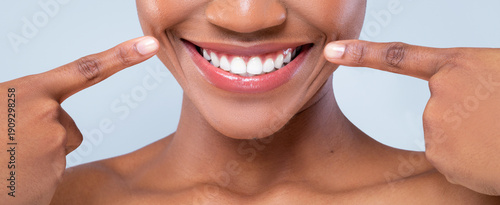 A woman is showing her bright smile while pointing at her teeth with her fingers. She is indoors against a light background. Her joy and positivity are clear in her expression.