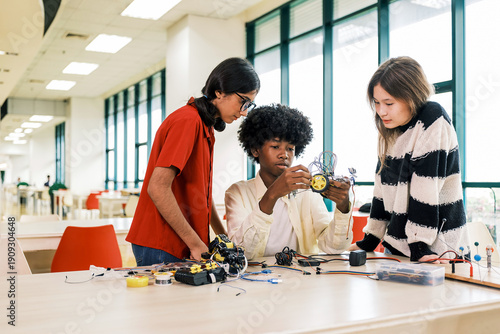 Three students collaborate to assemble a small wheeled robot in a bright classroom, surrounded by electronic components and tools.