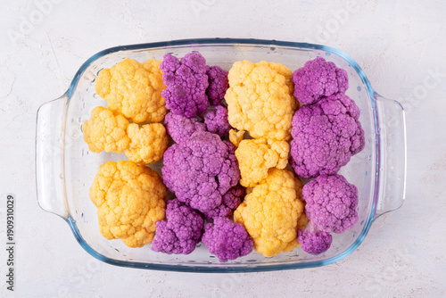 Yellow and purple cauliflower florets in a glass bowl on a white table. Top view. Preparing the ingredient