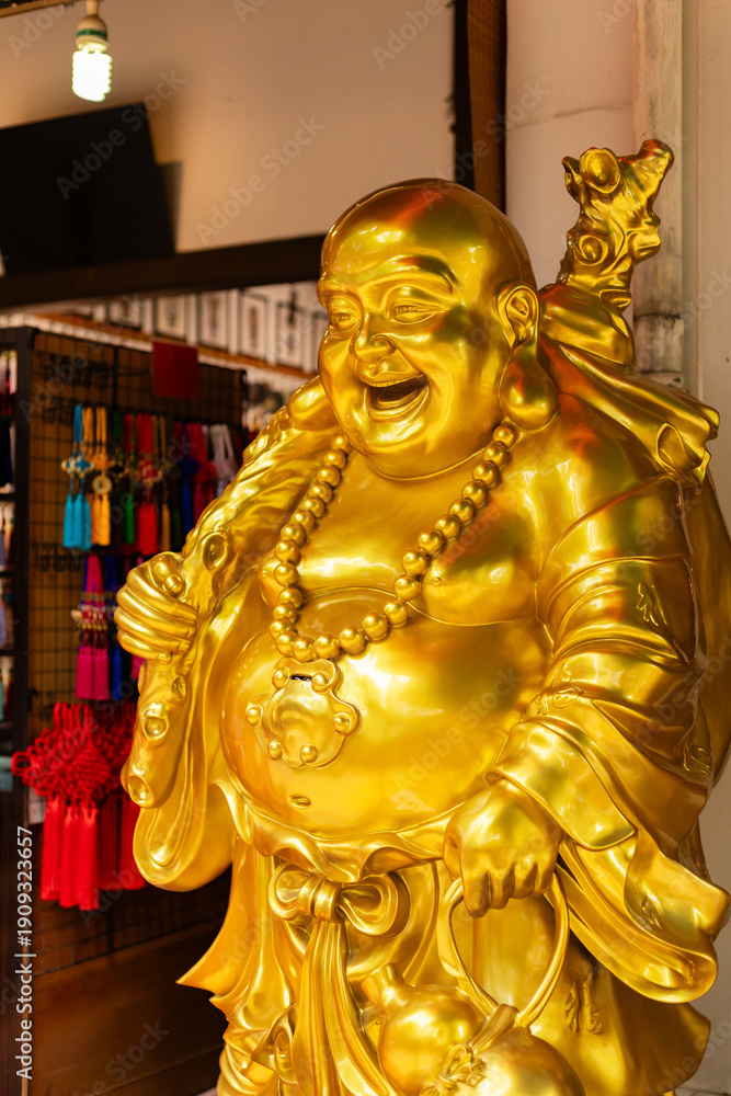 Fototapeta premium A golden laughing Buddha statue stands prominently inside a shop. The statue wears a large necklace and holds a cane. Colorful items hang in the background, showing a lively market scene