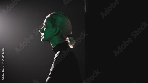 A young blonde woman with a short ponytail makes a step to the left, entering a beam of light. High-contrast side lighting sculpts her pensive profile, capturing the essence of overcoming silence and 