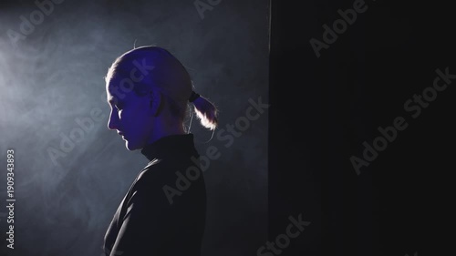 A young blonde woman with a ponytail takes a left step into a dramatic side light. High-contrast video emphasizes her contemplative profile, representing the moment of finding one's voice and facing t