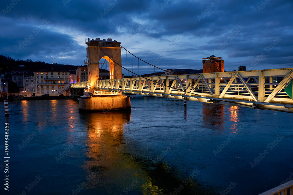 Fototapeta premium Illuminated pedestrian bridge over the Rhône river at night in Vienne, France. Beautiful night view of the Saint-Romain-en-Gal footbridge with golden lights reflecting in the water.