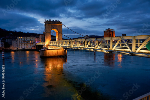 Illuminated pedestrian bridge over the Rhône river at night in Vienne, France. Beautiful night view of the Saint-Romain-en-Gal footbridge with golden lights reflecting in the water.