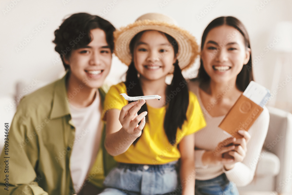 Fototapeta premium Smiling young girl in hat hold plane, selective focus. Millennial japanese parents happy and show passport and tickets in living room interior, blurred. Family vacation, travel and holiday together