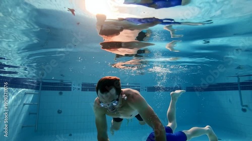 Father and child swim underwater in pool wearing goggles, surrounded by colorful toy fish on tiled floor. Concept of family fun, swimming lessons, and playful bonding in water environment.
