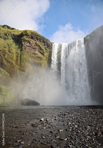 Skogafoss waterfall in southern Iceland with powerful water curtain, drifting mist and a rocky riverbed in the foreground under partly cloudy sky.