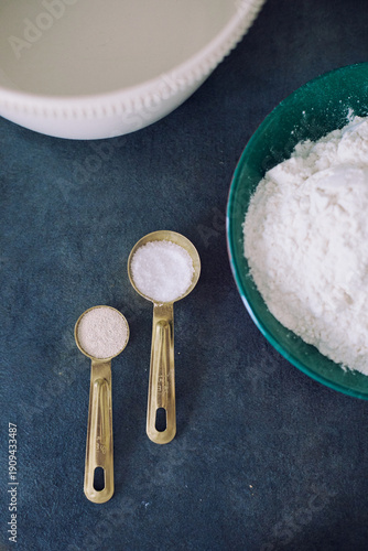Close-up of measuring spoons with salt and dry yeast beside a flour bowl on dark background. Ideal for culinary, food photography, and baking projects.