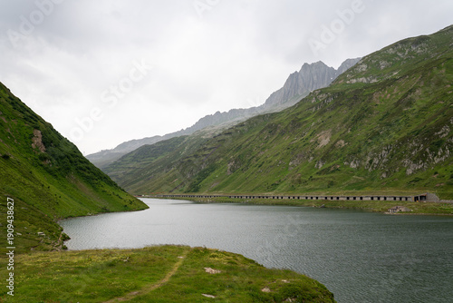 Bilde på lerret Serene mountain lake surrounded by lush green slopes and rugged peaks under an overcast sky, showcasing a calm alpine landscape and wide open scenery