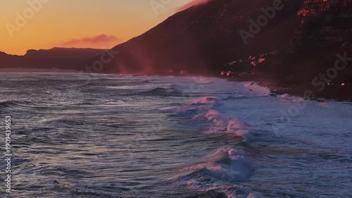 Aerial view of waves crashing on the coastline, with dramatic mountains in the background under a vibrant, colorful dawn sky, Cape Town, Western Cape, South Africa.