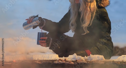Φωτογραφία Sitting beside a crackling campfire, a woman in a warm jacket pours hot chocolate into a metal mug