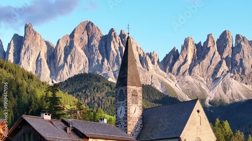 Aerial view of a church with a tall steeple, nestled in a valley with jagged mountains in the background, Santa Magdalena, Trentino-Alto Adige, Italy.
